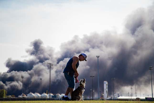 Un homme en short et casquette promène son chien devant un énorme panache de fumée noire